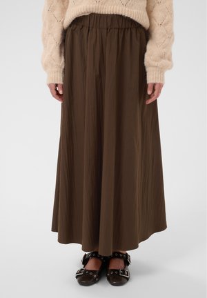 Personne portant un pull beige, une jupe marron longue jusqu’aux chevilles et des chaussures noires à boucles, debout devant un fond uni.