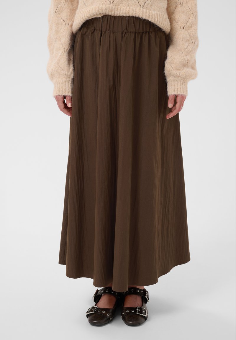 Personne portant un pull beige, une jupe marron longue jusqu’aux chevilles et des chaussures noires à boucles, debout devant un fond uni.