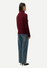 Femme de profil portant un pull à col roulé bordeaux, un jean bleu ample et des escarpins noirs à bouts pointus sur un fond uni.