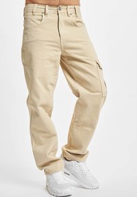 Beige cargo pants with a relaxed fit, made of cotton, featuring side pockets and a button closure. Paired with white sneakers.