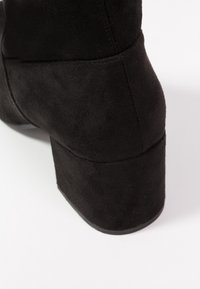 Czarny zamszowy botek za kostkę z masywnym obcasem typu blok, zaokrąglonym kształtem i subtelnym przeszyciem na zewnętrznej stronie.