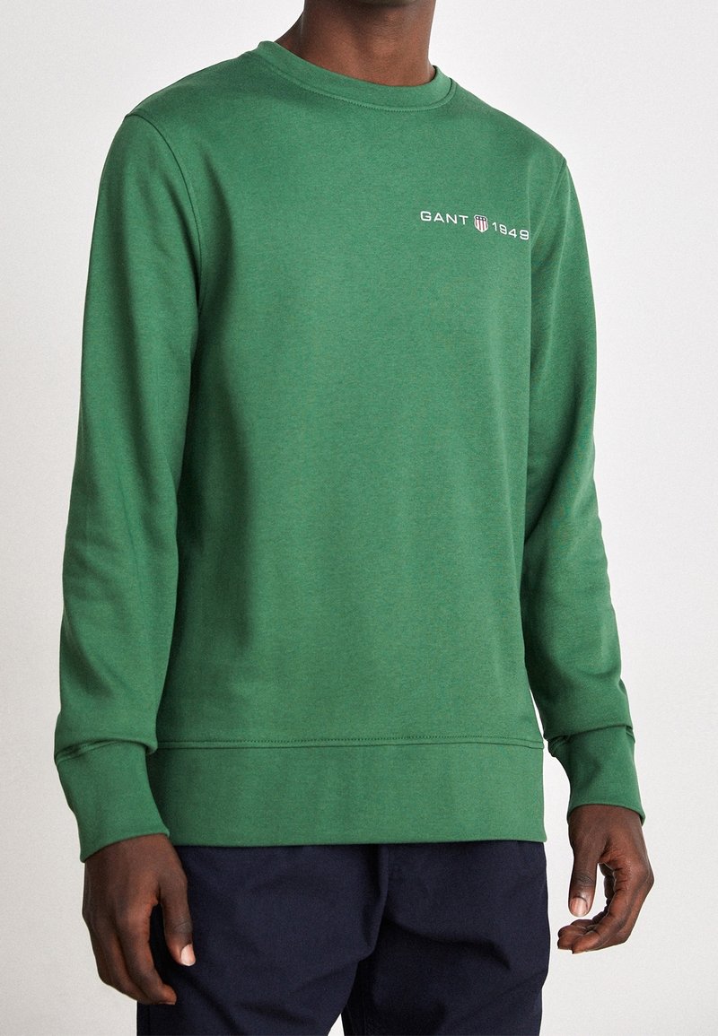 Personne portant un sweat-shirt vert à manches longues avec le texte « GANT 1949 » sur la poitrine, associé à un pantalon foncé.