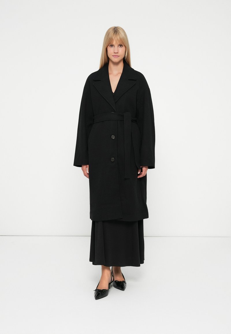 Mulher com cabelo loiro liso, vestindo um casaco preto com cinto sobre uma saia longa preta e sapatos pretos de bico afiado, em pé sobre fundo branco.