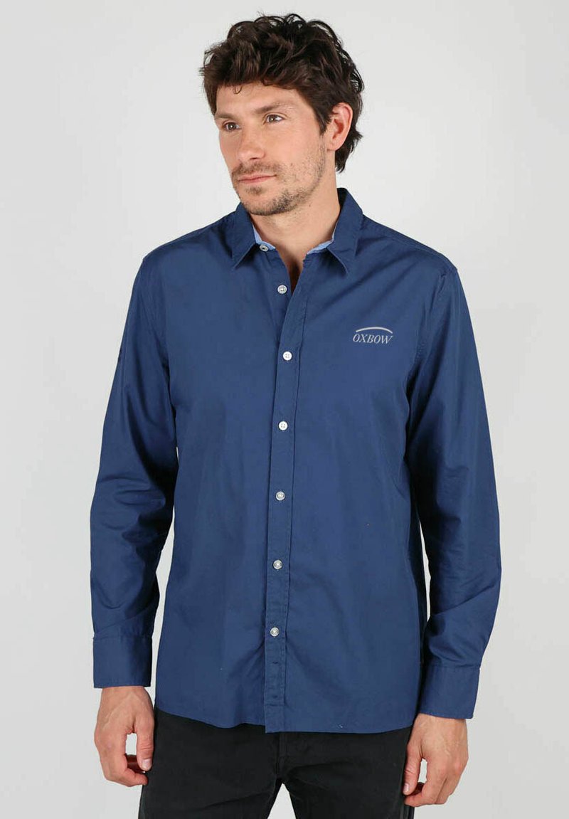 Chemise boutonnée bleu marine, manches longues, en tissu de coton, avec un petit logo sur la poitrine gauche, boutons blancs au toucher lisse.