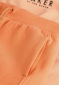 Tejido naranja claro, con una cinturilla acanalada y una textura suave, además de un detalle de bolsillo fruncido. Logo visible en negro en la parte delantera.