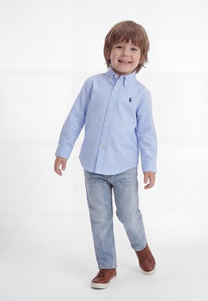 Χαμογελαστό νεαρό αγόρι περπατάει, φοράει ανοιχτό μπλε πουκάμισο με κουμπιά, ανοιχτό μπλε τζιν και καφέ αθλητικά παπούτσια σε απλό λευκό φόντο.