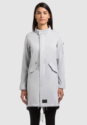 Femeie cu păr lung și închis la culoare, purtând o haină gri deschis până la mijlocul coapselor, cu buzunare cu clapetă și guler în picioare, stând în fața unui fundal simplu.