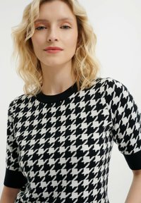 Kardigan v černobílém houndstooth vzoru s krátkými rukávy, černým vroubkovaným výstřihem a měkkou texturou.