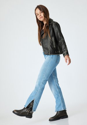 Zwart leren motorjack, lichtblauwe rechte jeans en zwarte enkellaarsjes met gespen. Model poseert voor een neutrale achtergrond.
