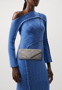Vestido azul acanalado con diseño off-shoulder y mangas largas. Clutch sobre con textura y patrón ondulado en color gris, llevado cruzado.