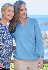 Due donne stanno insieme all'aperto. Una indossa una maglietta henley blu a maniche lunghe. L'altra ha un top a maniche corte con un motivo e tonalità di viola.
