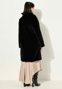 Abrigo de piel sintética negra con cuello, mangas largas y corte recto, combinado con una falda beis en capas y botas negras hasta la rodilla.