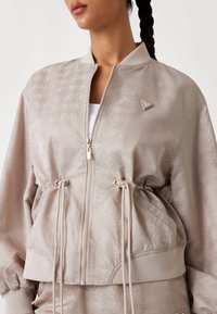 Light beige bomber jacket with a textured pattern, drawstring waist, ribbed cuffs, and zip closure, featuring a small triangular logo accent.