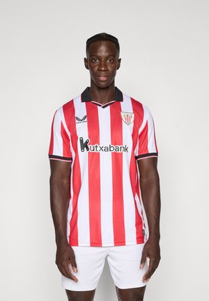 Man in een rood en wit gestreepte Atletiek Club Bilbao voetbalshirt en witte shorts, staande tegen een effen lichte achtergrond.