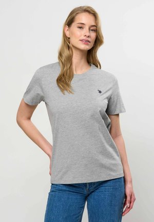 Kvinde med langt blondt hår iført en lysegrå T-shirt med korte ærmer og blå jeans, stående mod en ensartet lys baggrund.