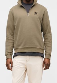 Homme portant un sweat-shirt à col zippé olive par-dessus une chemise sombre et un pantalon beige clair, debout devant un fond uni.