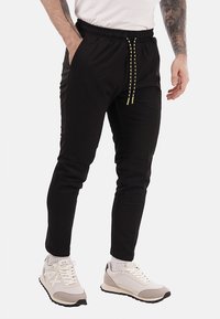 Pantalones de chándal negros con cintura elástica y cordón decorativo amarillo. Diseño entallado, tela suave y bolsillos laterales. Combinados con zapatillas blancas.