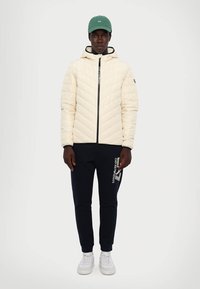 Lys beige quiltet jakke med hette, med en front glidelås. Parret med marineblå joggebukser og hvite sneakers, toppet med en grønn caps.