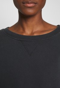 Musta collegepaita, jossa on resorinen pääntie, edessä V-ommel-yksityiskohta ja pehmeä, teksturoitu kangas. Minimalistinen muotoilu.