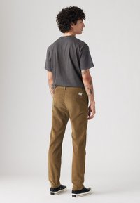 Pantalones de pana marrones con corte recto, combinados con una camiseta gris oscuro. Presentan una ligera textura y bolsillos traseros con una etiqueta. Los zapatos son negros.