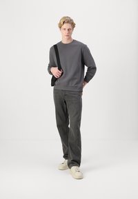 Gray sweatshirt with long sleeves, crew neckline; paired with gray, loose-fitting pants and white sneakers. Model holds a black bag.