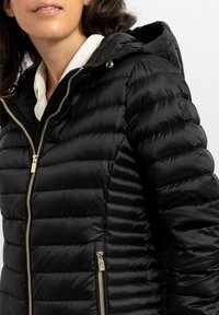 Giacca imbottita nera con cappuccio, caratterizzata da un design trapuntato, zip dorate e una toppa con logo sulla manica. Texture liscia e lucida.