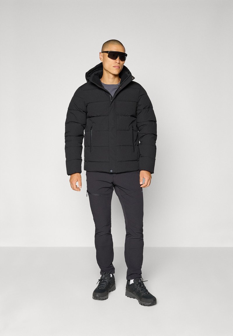 Schwarze Pufferjacke mit Kapuze, gestepptem Design, zwei Reißverschluss-Taschen, kombiniert mit schwarzen Hosen und robusten schwarzen Schuhen. Zubehör umfasst eine Sonnenbrille.