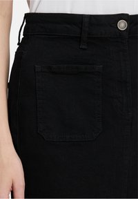 Gros plan sur une jupe en denim noir montrant la poche arrière, la taille avec bouton, la passante de ceinture et une partie de la main et du torse d'une personne.
