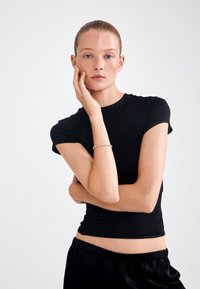 Melns piegriezts T-krekls ar īsām piedurknēm, gluda tekstūra. Valkāts kopā ar brīviem melniem biksēm. Modelis ar neitrālu izteiksmi, roka uz sejas.