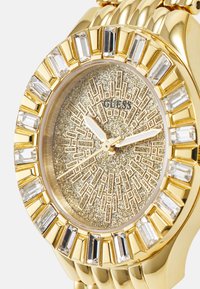 Goldfarbene Uhr mit einem funkelnden, goldtexturierten Zifferblatt, rechteckigen Kristallen um die Lünette und schlanken Stunden- und Minutenzeigern.