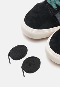 Black suede sneakers with white rubber sole, featuring teal laces. Two black round shoelaces placed next to the sneakers.