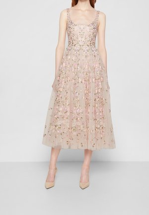 Femme portant une robe midi en tulle beige clair sans manches avec une broderie florale rose et verte, associée à des escarpins beiges à bout pointu.