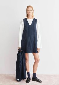 Marineblauwe jurk met krijtstrepen en een V-hals, gecombineerd met een wit overhemd met lange mouwen. Model houdt een bijpassend jasje vast. Zwarte schoenen en donkere sokken.