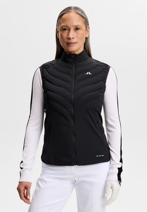 Frau trägt schwarze gesteppt Weste über weißem Langarmshirt mit schwarzen Streifen, weiße Hose und weißen Golfhandschuh an der linken Hand.