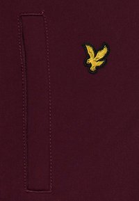 Burgundy tyg med en strukturerad yta som har en liten gul broderad örnlogga på vänster sida och en sydd fickdetalj.
