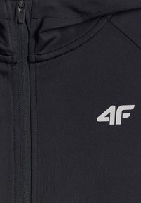 Veste noire zippée avec fermeture éclair visible et logo blanc "4F" sur la poitrine, montrant la texture du tissu et les détails des coutures.
