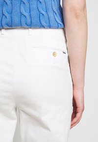 Bílé kalhoty s hladkou texturou, s kapsou na zadní straně zdobenou dřevěným knoflíkem. Nošeny s modrým svetr z pleteniny s copánkovým vzorem.