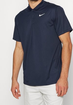 Mand iført en mørkeblå Nike polo med korte ærmer og hvidt logo på brystet samt hvide bukser, vist fra midt på brystet til hofterne.