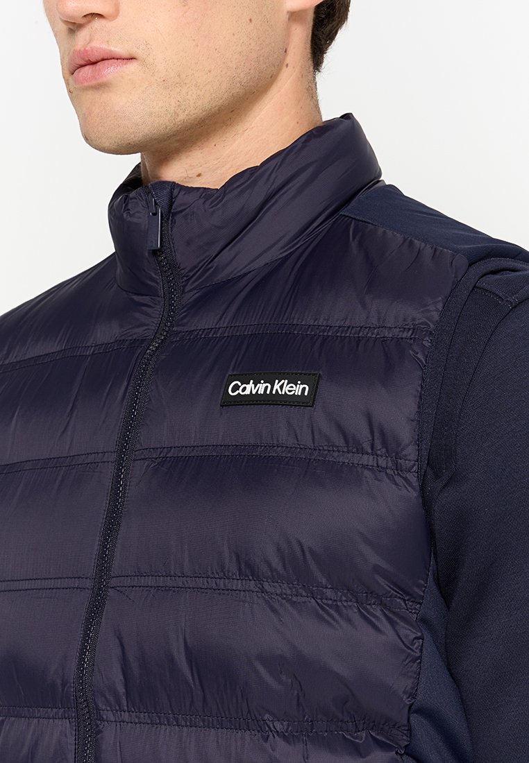 Gilet matelassé bleu marine avec un col montant, doté d'une fermeture éclair à l'avant et d'une texture matelassée horizontale. Logo Calvin Klein noir sur la poitrine.