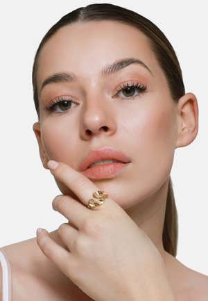 Nahaufnahme einer Frau mit natürlichem Make-up, die ihr Kinn berührt, trägt einen goldenen Statement-Ring, mit zurückgebundenen Haaren vor weißem Hintergrund.