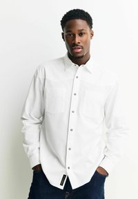 Hvid skjorte med knapper og struktureret stof, to brystlommer og sølvknapper. Bæres med mørke denim jeans. Etiket synlig ved nederste kant.