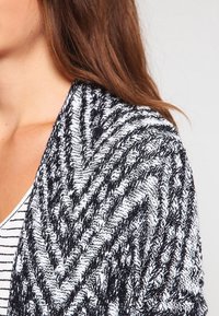 Châle tricoté en noir et blanc avec un motif en zigzag, présentant un fil texturé. Design doux et fluide qui tombe sur les épaules.