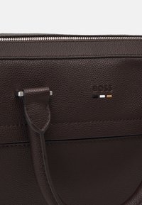 Tummanruskea teksturoitu nahkakassi, jossa on kahva, vetoketju ja pieni kohokuvioitu "BOSS"-logo, jonka alla on mustia, valkoisia ja hiekansävyisiä raitoja.