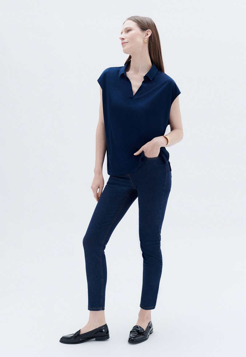 Blusa blu navy a maniche corte con colletto a punta, abbinata a jeans skinny blu scuro. Il modello indossa mocassini neri e gioielli minimal in oro.