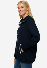 Giacca zip-up blu navy con colletto alto, polsini a costine e due tasche laterali che presentano accenti bianchi e un tessuto morbido e strutturato.
