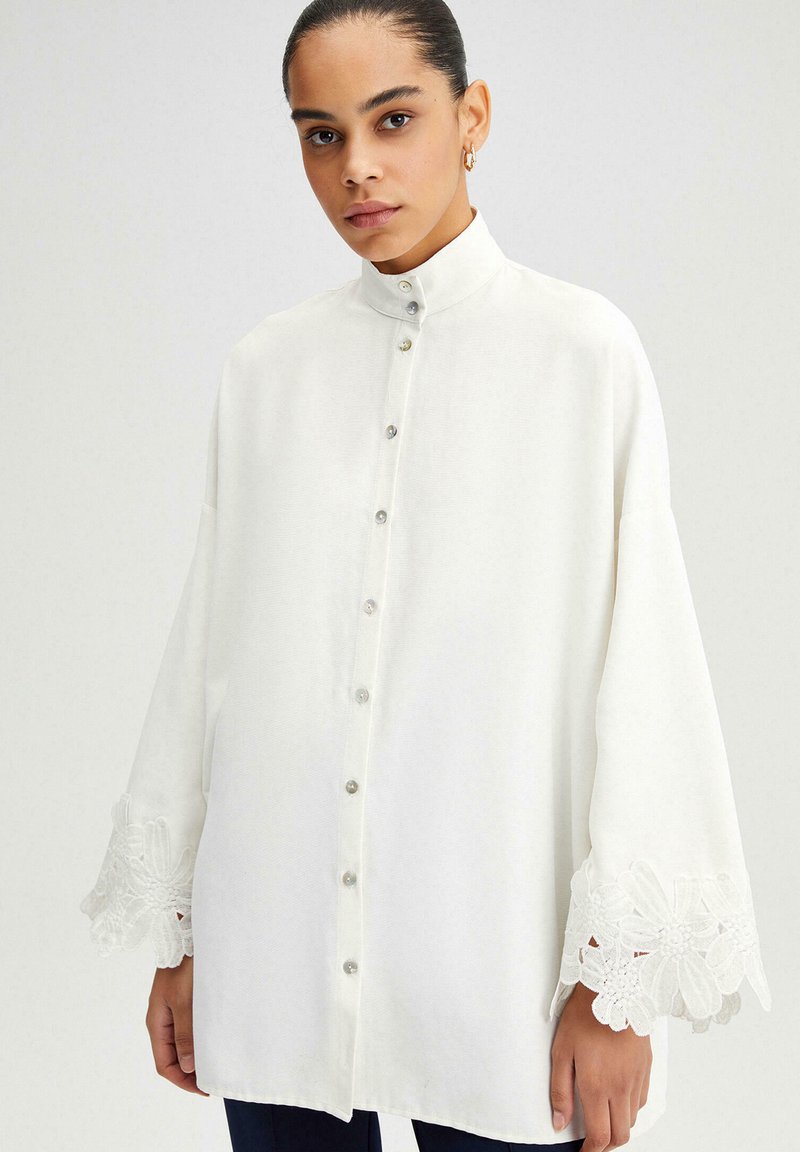 Femme portant une blouse blanche ample à boutons avec un col montant et des détails en dentelle florale sur les manches larges, debout devant un fond uni.