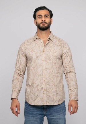 Homme aux cheveux foncés et à la barbe, portant une chemise beige à manches longues à motifs et un jean bleu, debout devant un fond clair uni.