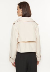 Lichtbeige cropped jack met een brede kraag, knoopsluitingen aan de voorkant en knopen op de mouwen. Gladde stof met een ventilerend detail aan de achterkant.