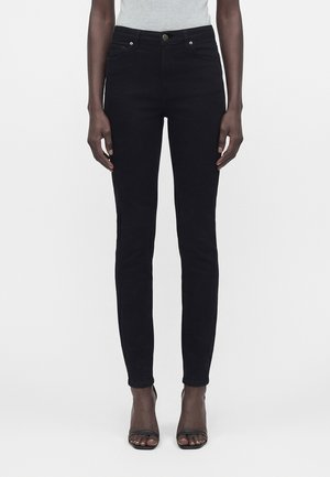 Kobieta w dopasowanych czarnych spodniach i czarnych butach na otwartych obcasach, stojąca na jednolitym białym tle.