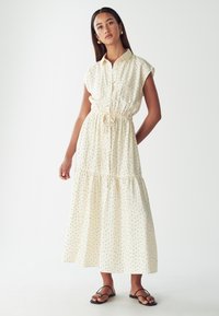 Svetle béžové maxi šaty s černými puntíky, krátkými rukávy, knoflíkovou přední částí, elastickým pasem a volánkovou sukní, doplněné černými sandály.